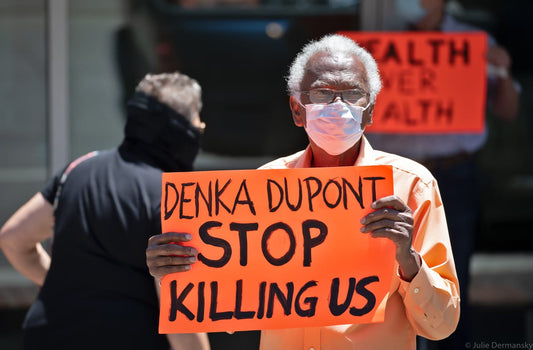 Protester wearing a mask holds a bright orange sign reading “Denka Dupont Stop Killing Us,” highlighting the health impacts of chloroprene pollution.