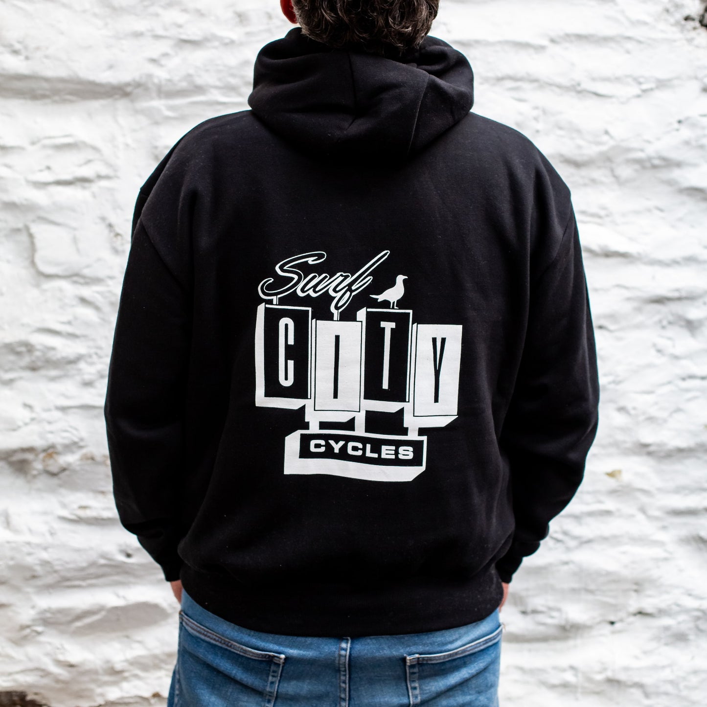 Person wearing a black hoodie with 'Surf City Cycles' logo on the back against a textured white wall at Project 83.