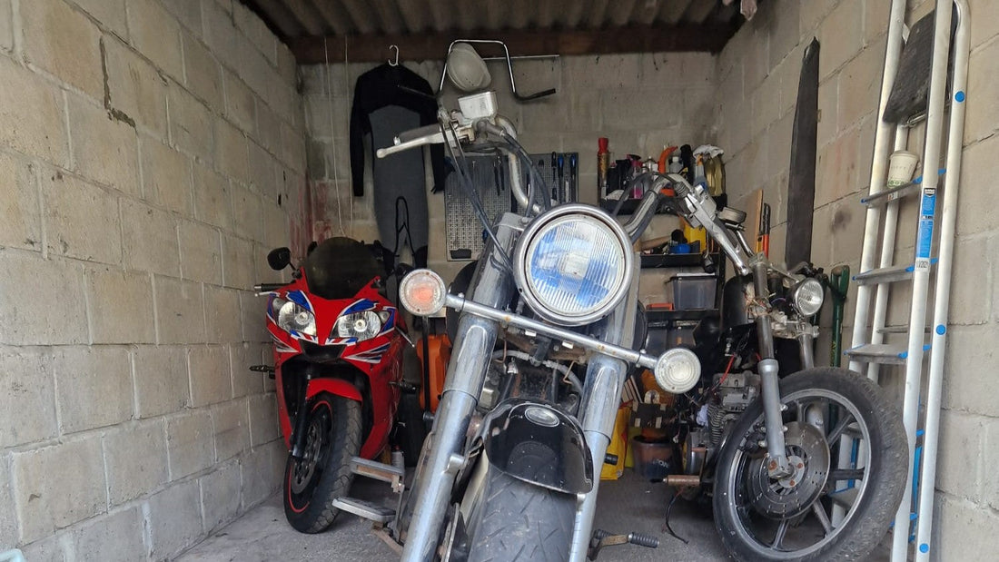 A garage with three motorcycles, including a red sportbike, a cruiser with a large round headlight in the foreground, and a partially disassembled bike. Tools, a wetsuit, and a ladder are stored against the concrete block walls.