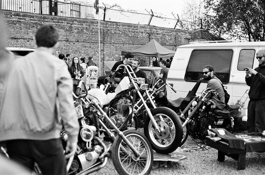 A black-and-white photo of a motorcycle gathering, featuring custom choppers, riders, and a lively crowd near a van and market stalls in an urban setting