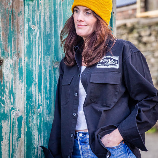 Woman wearing a black jacket with a logo, yellow beanie, and blue jeans against a wooden wall.