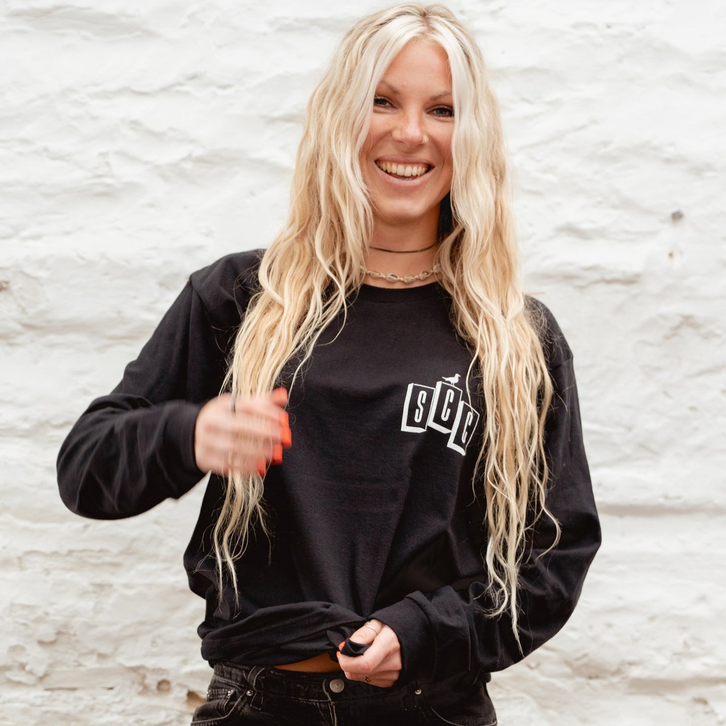 Lifestyle shot of a blonde surf girl wearing the black long sleeve Surf City Cycles OG logo tee, capturing a relaxed, rugged aesthetic.