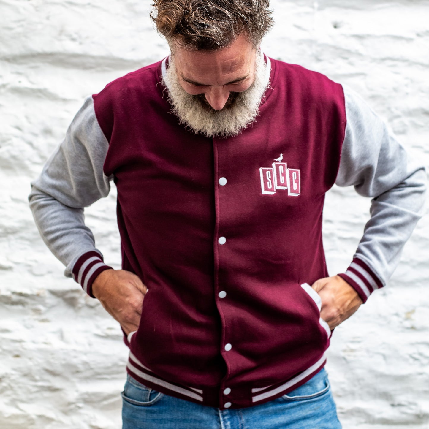 Bearded man wearing a burgundy and heather grey varsity jacket against a white stone wall.
