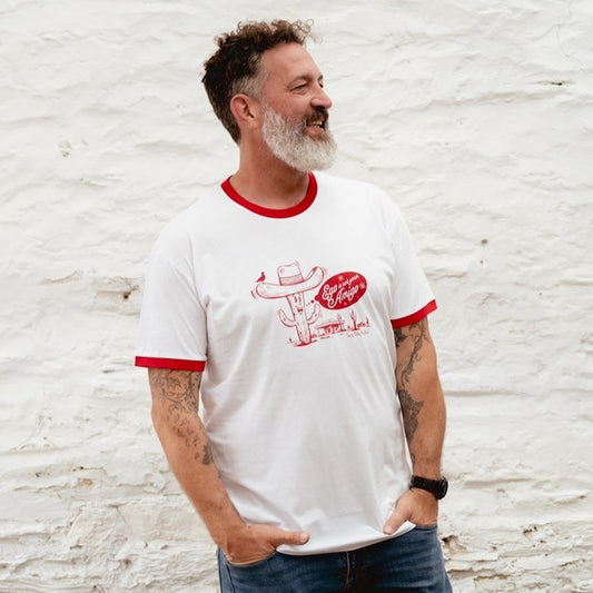 Bearded man wearing the white and red ringer tee with "Ego is not your Amigo" cactus graphic against a white stone wall.