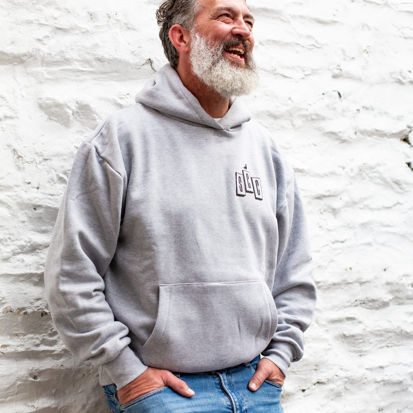 Bearded man wearing the heather grey Surf City Cycles OG Logo Heavyweight Hoodie with black branding against a stone wall.