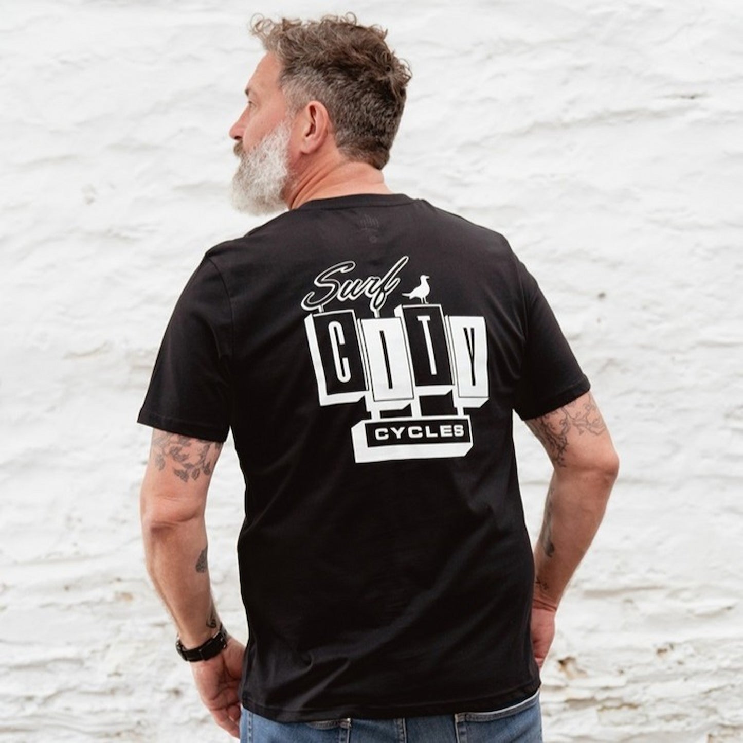 Back view of a male model looking over his shoulder wearing the Black OG Logo organic cotton tee, showcasing the large white Surf City Cycles branding.