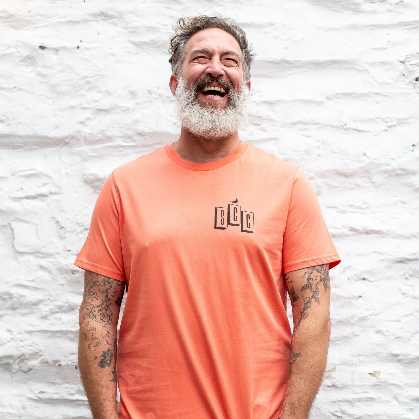 A male model with a grey beard laughing while wearing the Fiesta OG Logo organic cotton tee, showing the chest branding.