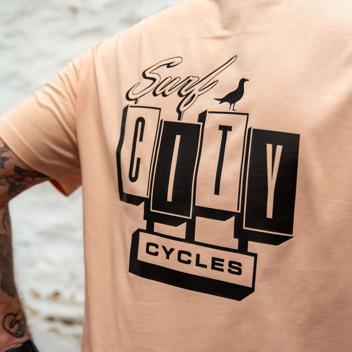 A detailed close-up of the back of the Peach OG Logo organic cotton tee, featuring the large Surf City Cycles block logo and bird graphic.