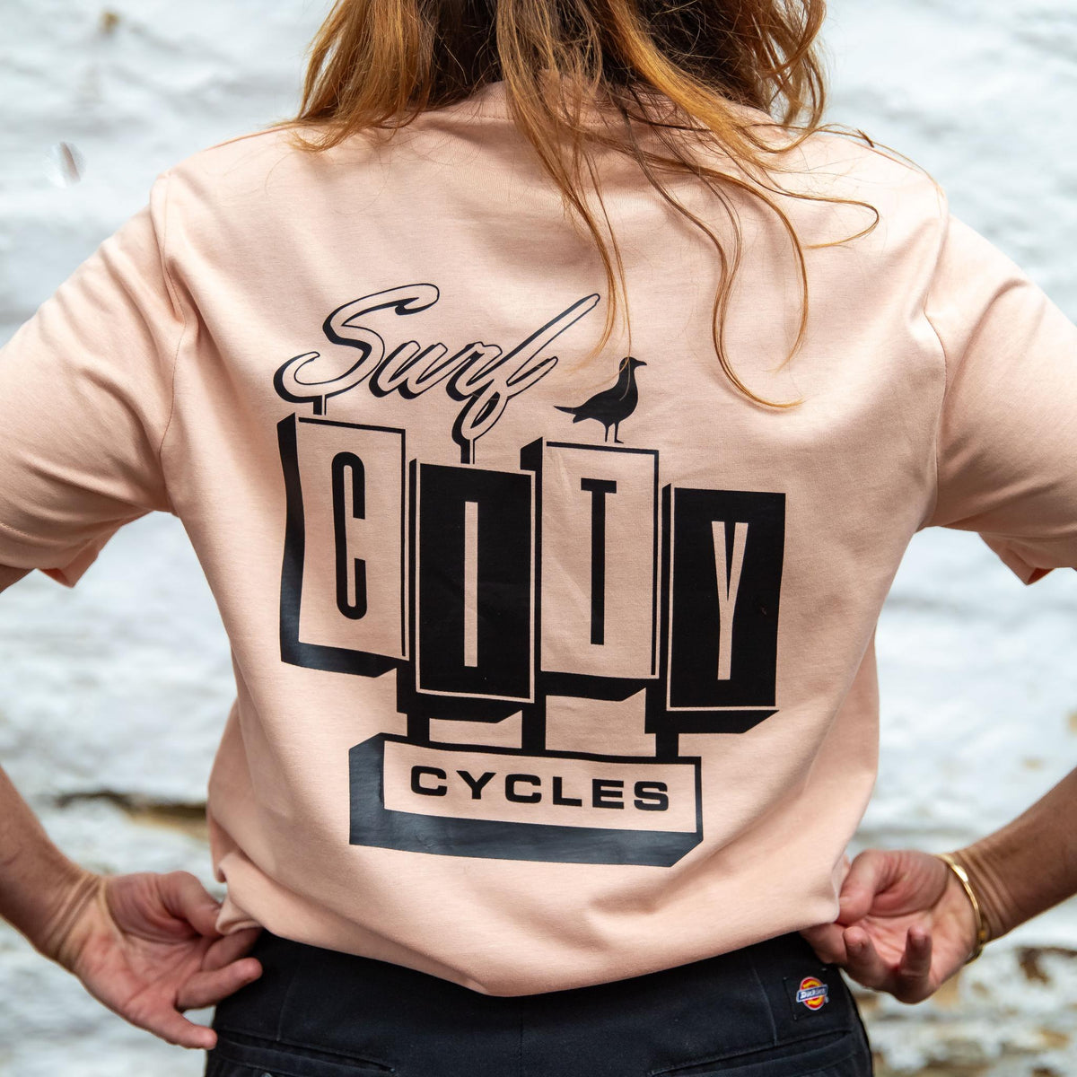 The back of the Peach OG Logo organic cotton tee worn with black trousers, showing the large Surf City Cycles block logo and bird graphic.