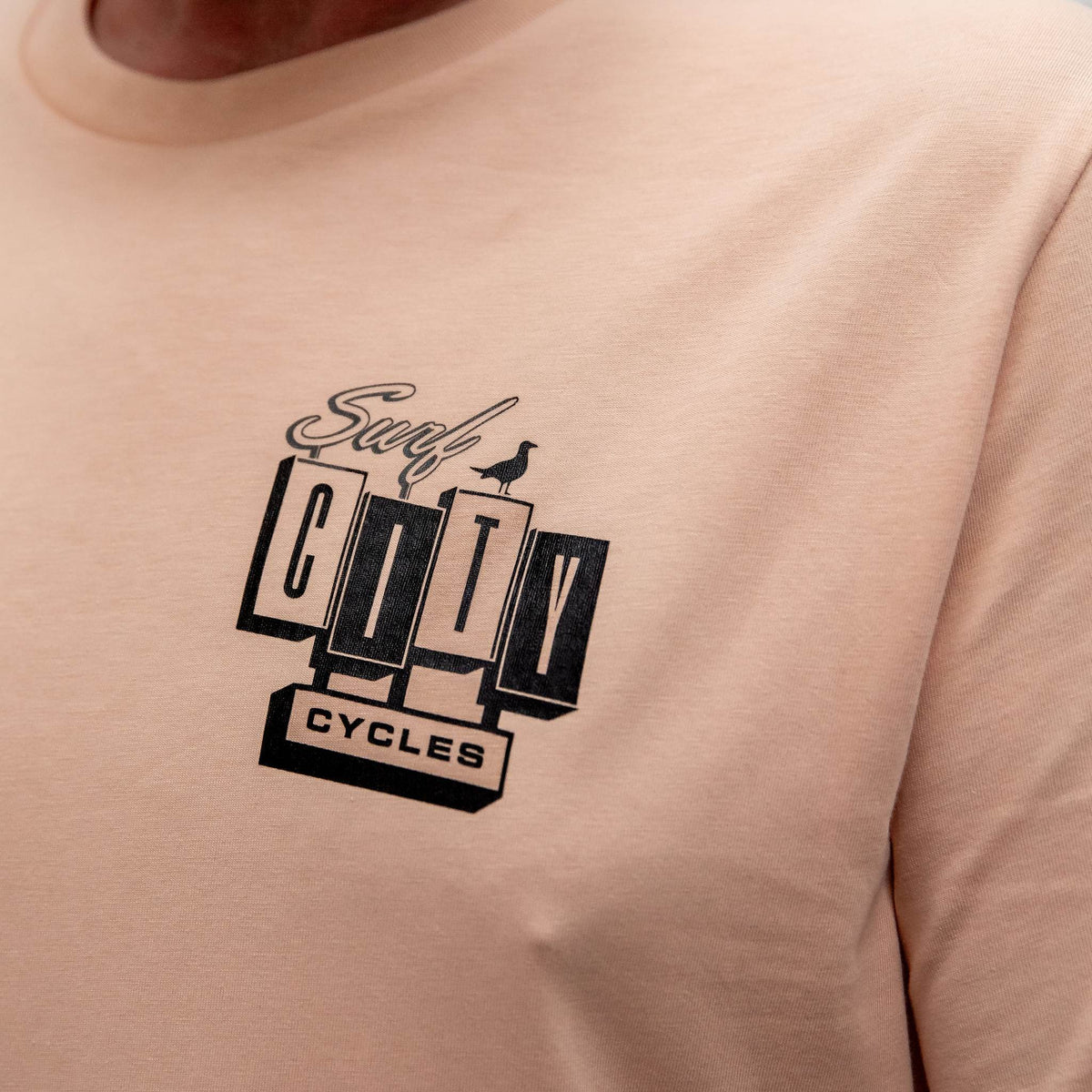 A detailed close-up of the front of the Peach OG Logo organic cotton tee, featuring the small Surf City Cycles block logo and bird graphic on the left chest.