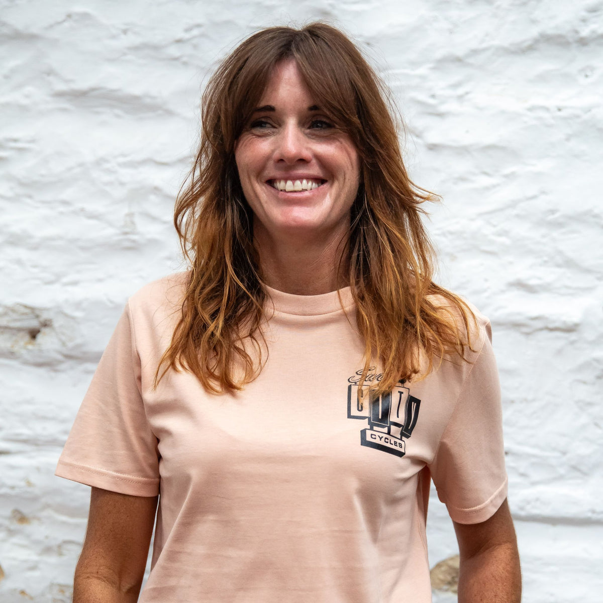 A woman smiling against a whitewashed stone wall, wearing the Peach OG Logo organic cotton tee with the small Surf City Cycles logo on the left chest.