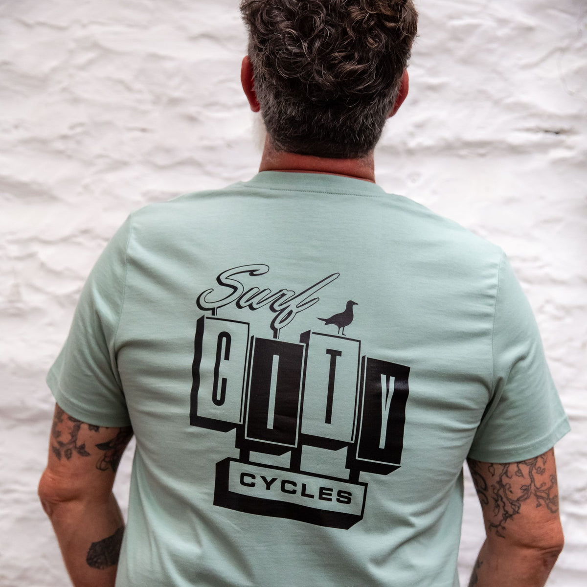 The back of the Sage OG Logo organic cotton tee worn by a man with tattooed arms, showing the large Surf City Cycles block logo and bird graphic.