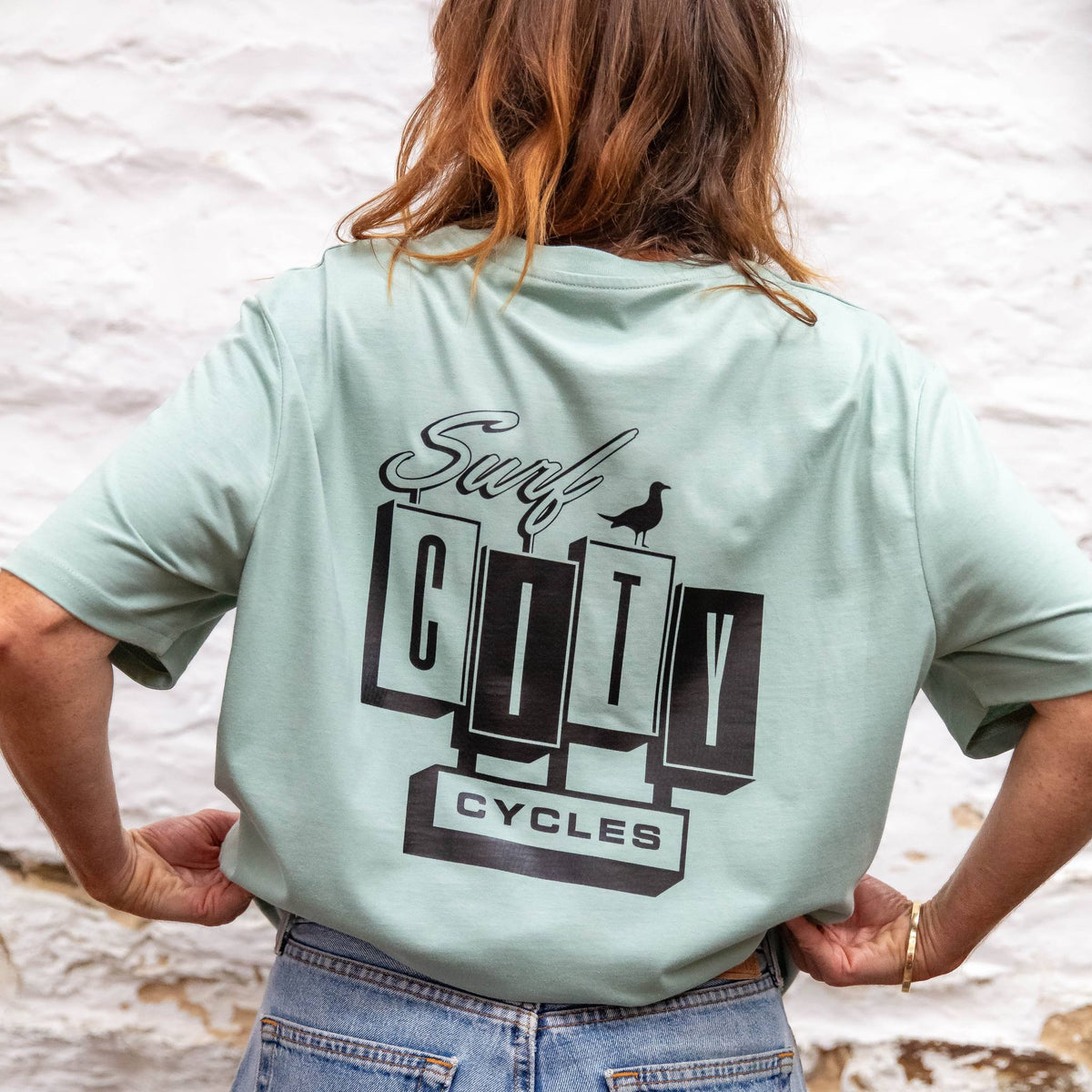 The back of the Sage OG Logo organic cotton tee worn with denim shorts, showing the large Surf City Cycles block logo and bird graphic.