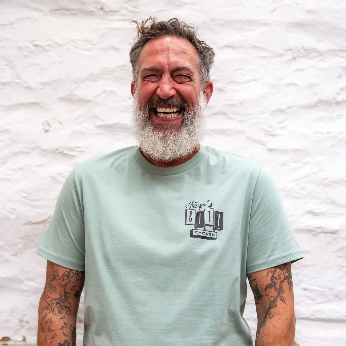 A bearded man with tattooed arms laughing against a whitewashed stone wall, wearing the Sage OG Logo organic cotton tee with the small Surf City Cycles logo on the left chest.