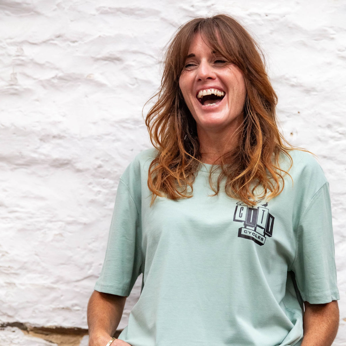 A woman laughing against a whitewashed stone wall, wearing the Sage OG Logo organic cotton tee with the small Surf City Cycles logo on the left chest.