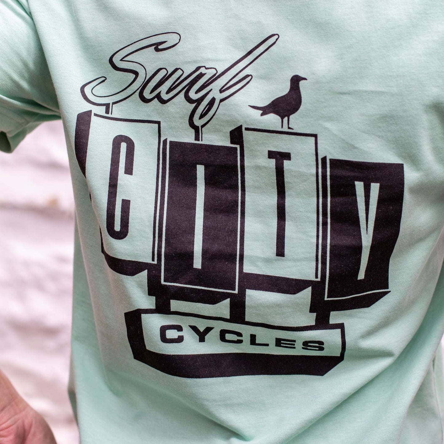 A detailed close-up of the back of the Seafoam OG Logo organic cotton tee, featuring the large Surf City Cycles block logo and bird graphic.