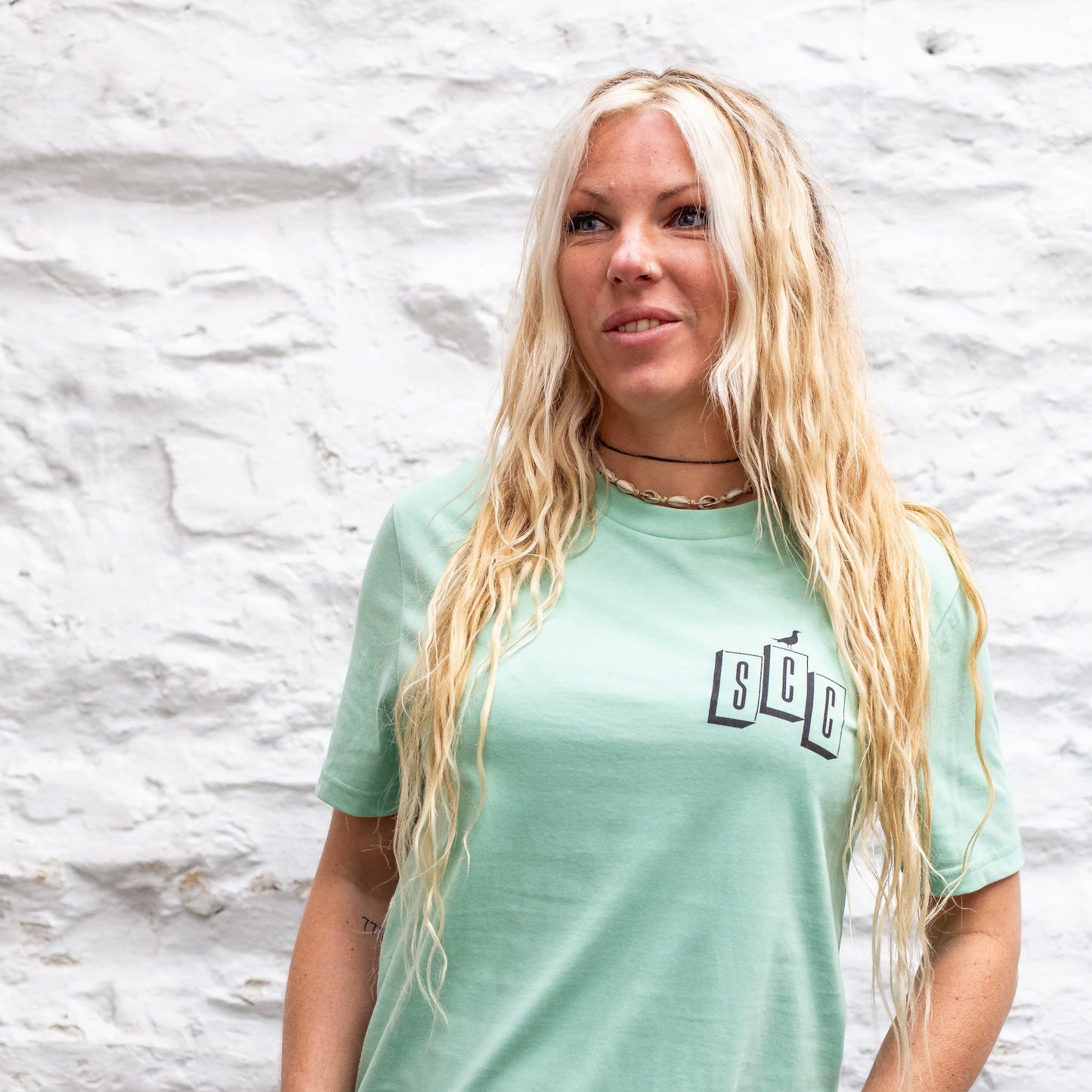 A female model wearing the Seafoam OG Logo organic cotton tee, featuring the SCC block logo on the chest, shot against a white stone wall.