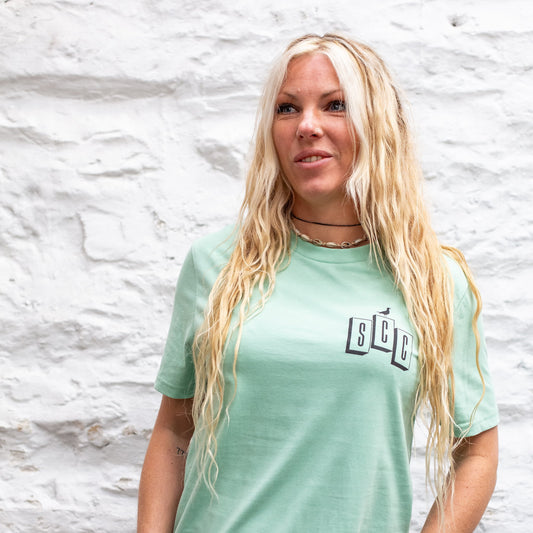 A female model wearing the Seafoam OG Logo organic cotton tee, featuring the SCC block logo on the chest, shot against a white stone wall.