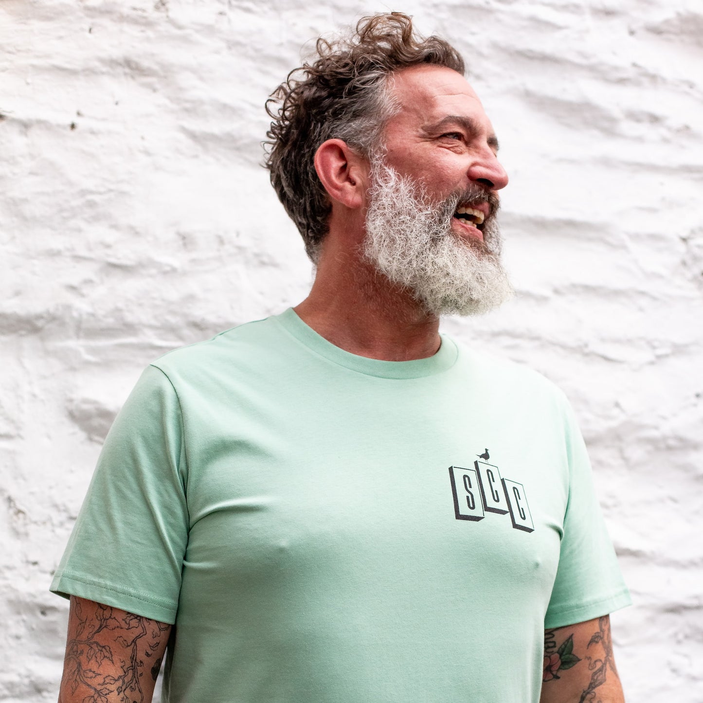 A male model with a grey beard laughing while wearing the Seafoam OG Logo organic cotton tee, showcasing the SCC block chest branding.