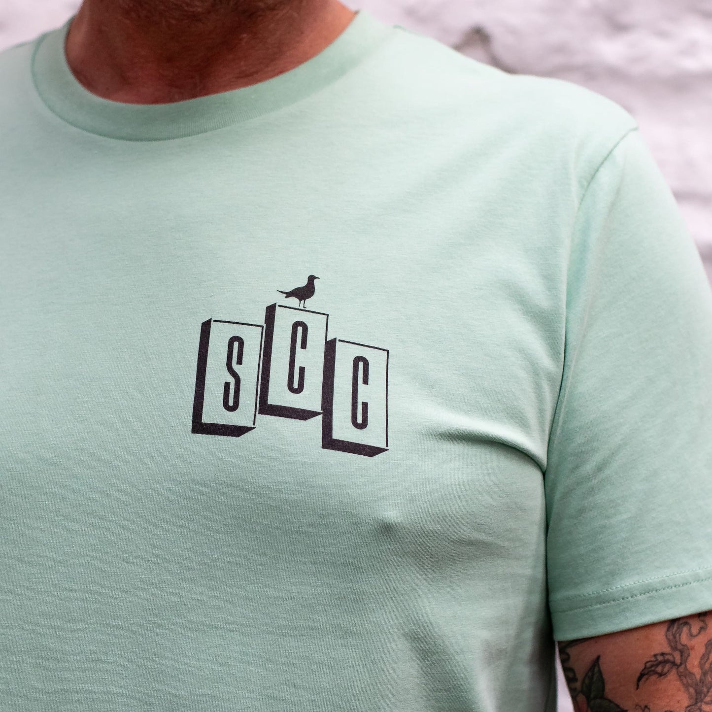 A detailed close-up of the SCC block logo and bird graphic on the chest of the Seafoam organic cotton tee.