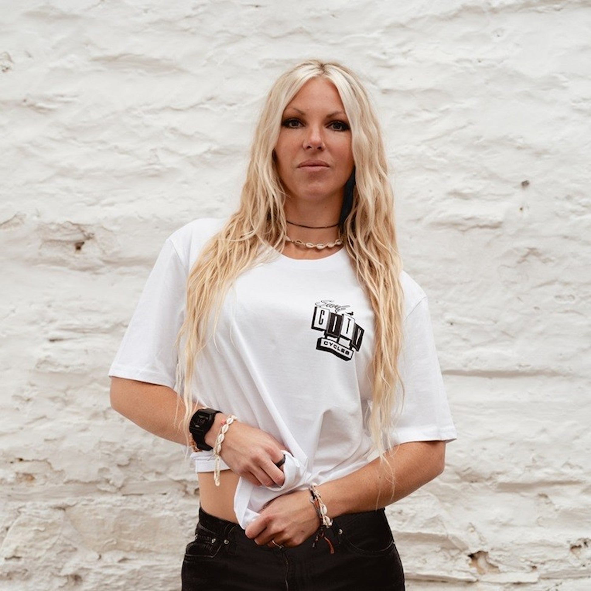 A female model wearing the OG Logo organic cotton tee in White, featuring the Surf City Cycles block logo on the chest, shot against a white stone wall.