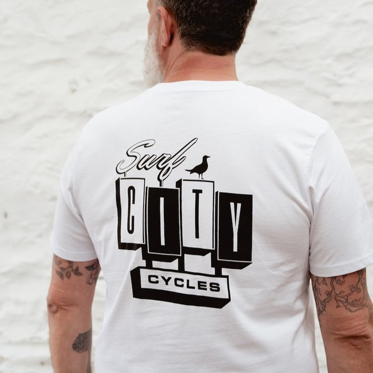 Back view of the male model wearing the white OG Logo organic cotton tee, showcasing the large Surf City Cycles block logo and bird graphic.