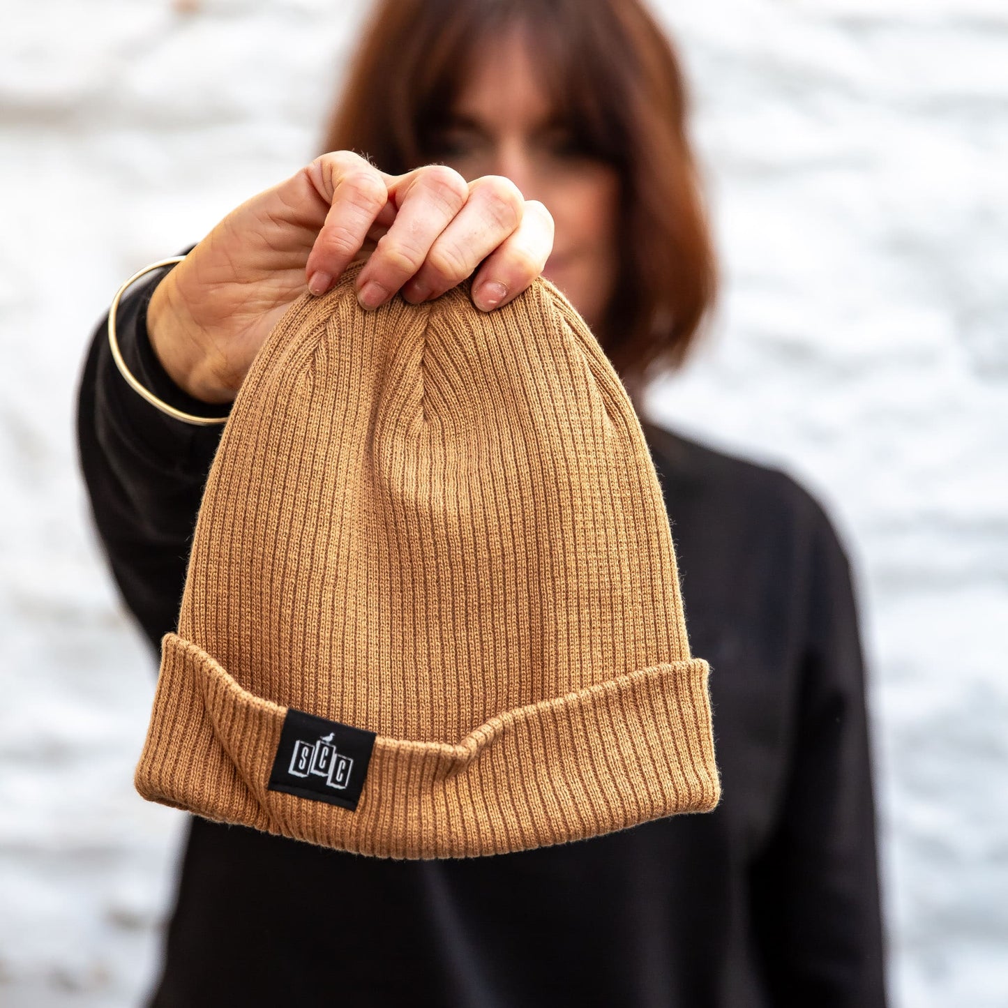 The Seafarer Organic Ribbed Beanie in Biscuit brown, held forward to show the SCC woven clip label detail.