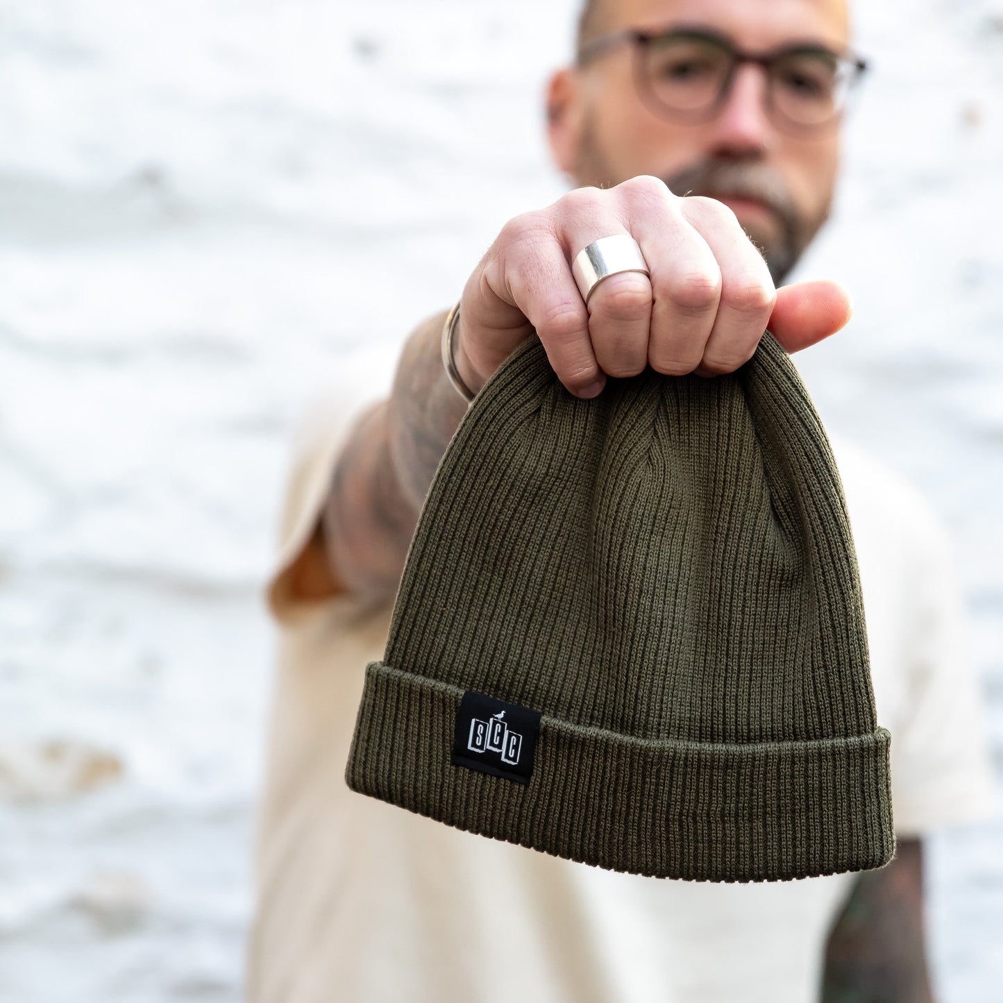 The Seafarer Organic Ribbed Beanie in Olive green, held forward to show the SCC woven clip label detail against a white stone wall.