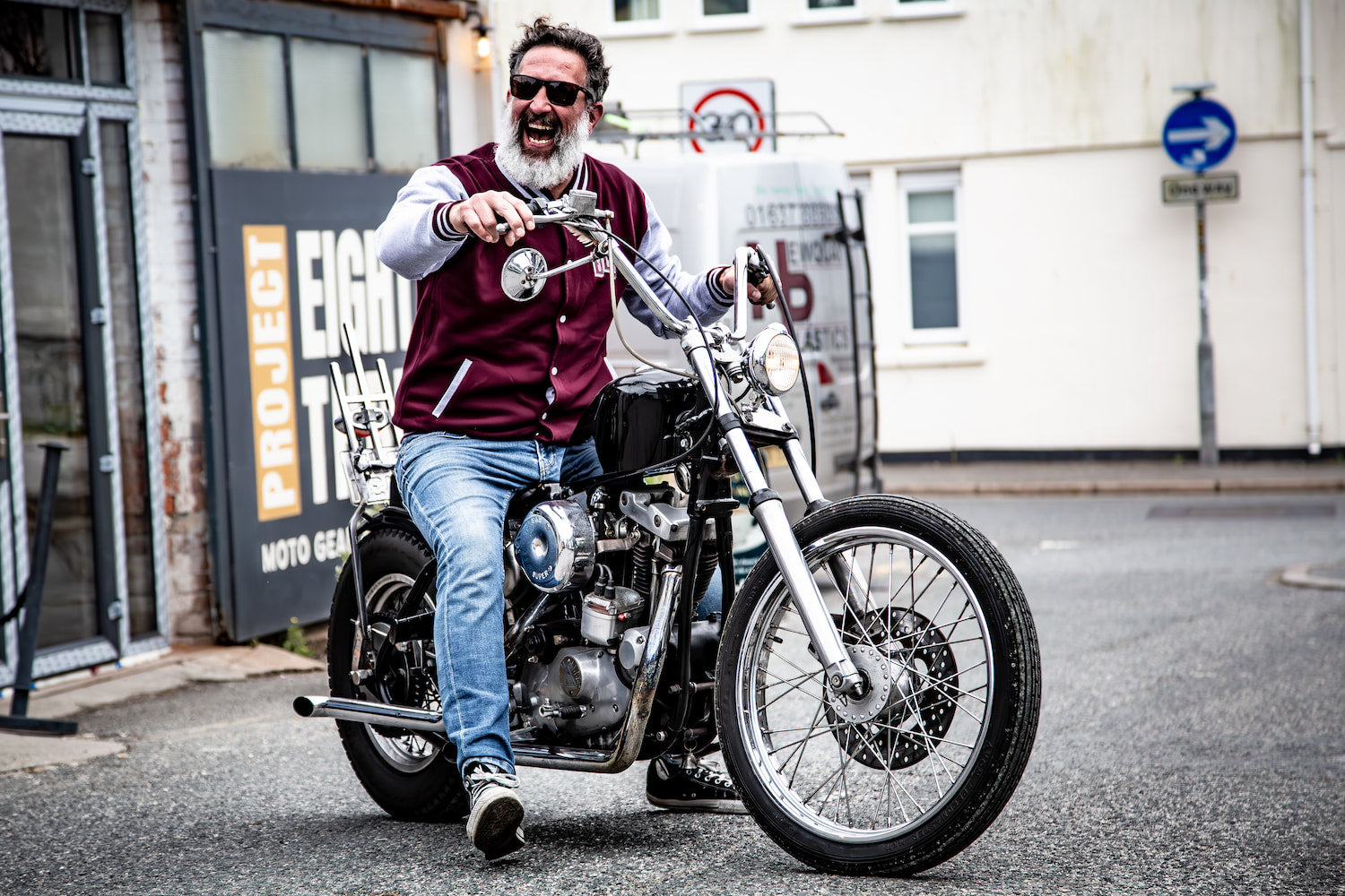 bearded man in a maroon varsity jacket laughing while sitting on a custom chrome chopper motorcycle outside Project 83 in Newquay.