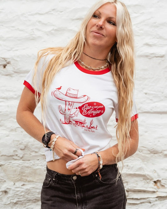 White ringer tee by Surf City Cycles with red neckline and sleeve cuffs, front view on female model, “Ego is Not Your Amigo” red cactus graphic, unisex