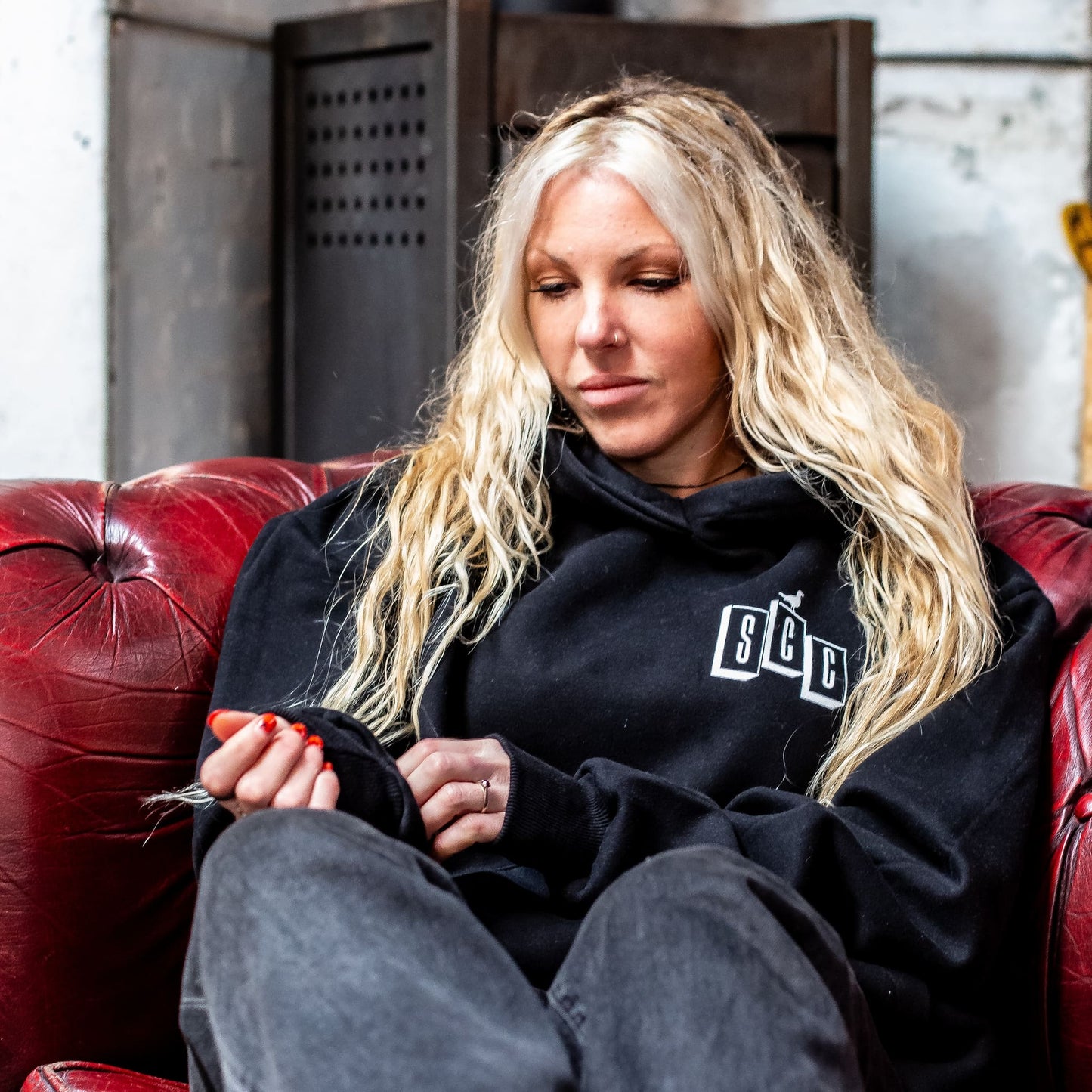 Blonde surf girl wearing a black Surf City Cycles hoodie sitting on a red leather couch in Project 83 Newquay