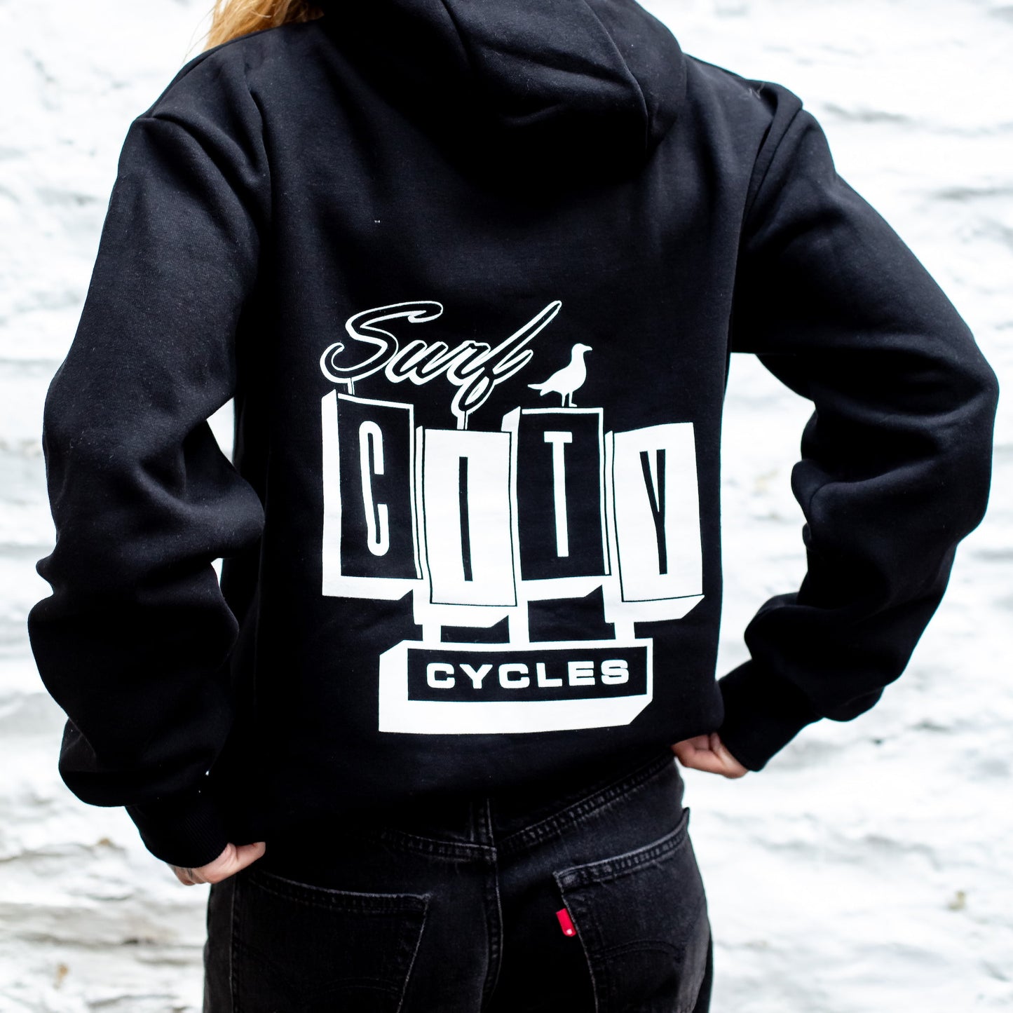 Person wearing a black hoodie with 'Surf City Cycles' logo on the back, standing against a blurred natural background.