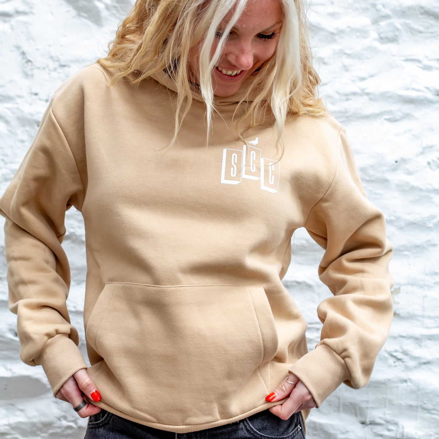 Front view of the Desert Sand Surf City Cycles heavyweight hoodie showing the black OG logo against a white stone wall in Project 83 Newquay.