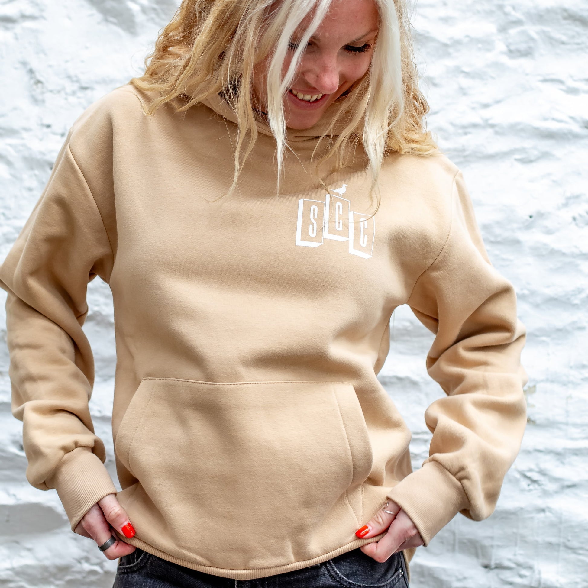 Front view of the Desert Sand Surf City Cycles heavyweight hoodie showing the black OG logo against a white stone wall in Project 83 Newquay.