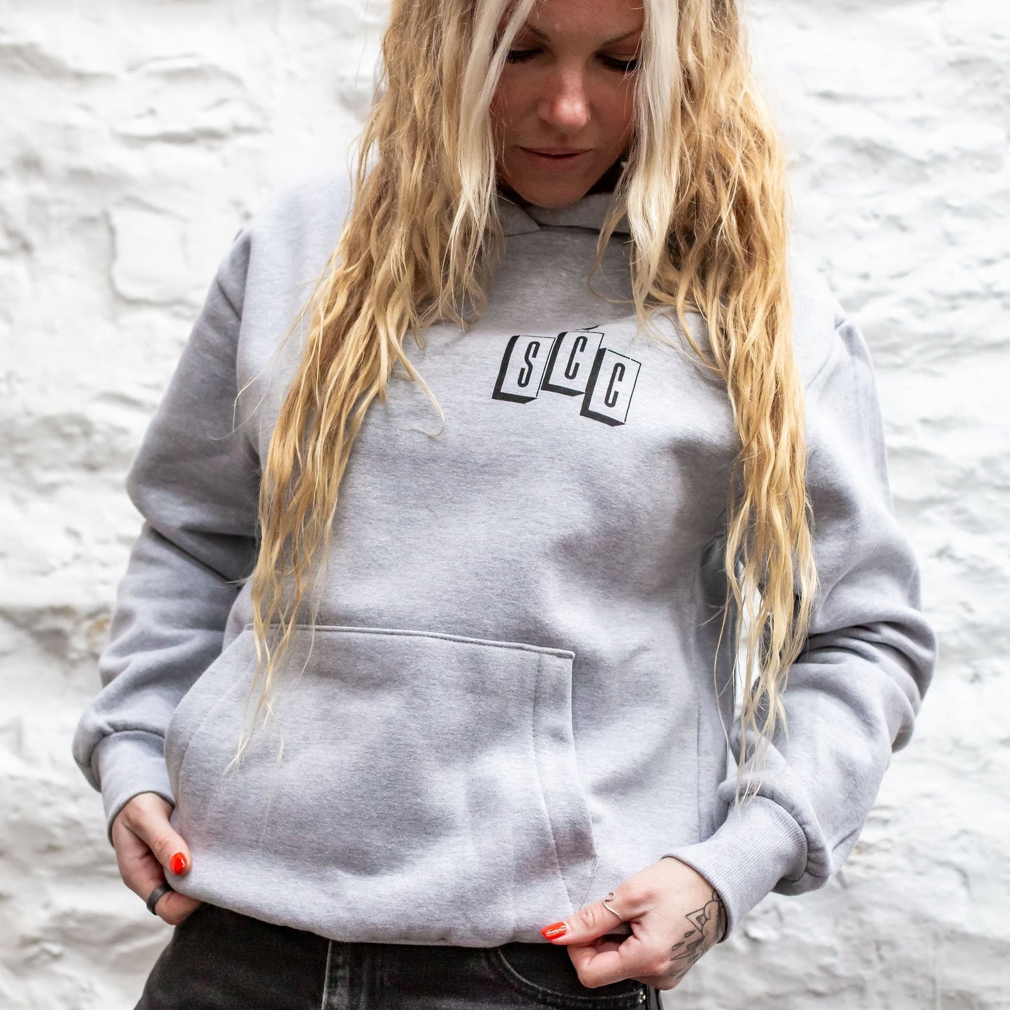 Female model wearing the heather grey OG Logo Heavyweight Hoodie with black chest branding against a white wall in Project 83 Newquay.