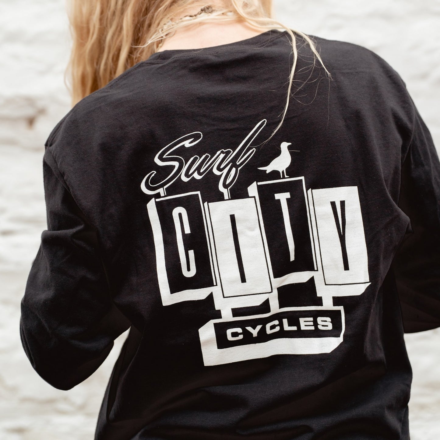 Medium shot of the Surf City Cycles black long sleeve OG logo tee against a white wall, showing the clean silhouette and ribbed cuffs.