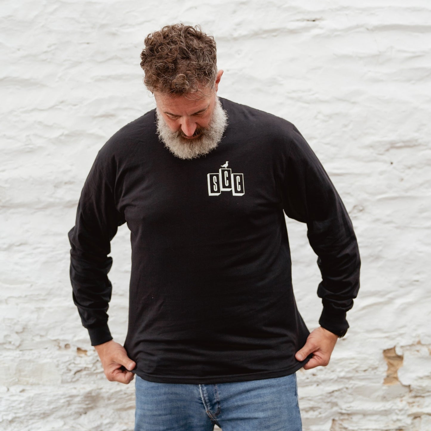 Andrew Pringle wearing the Surf City Cycles black long sleeve OG logo shirt, leaning against a wall in Project 83 Newquay.