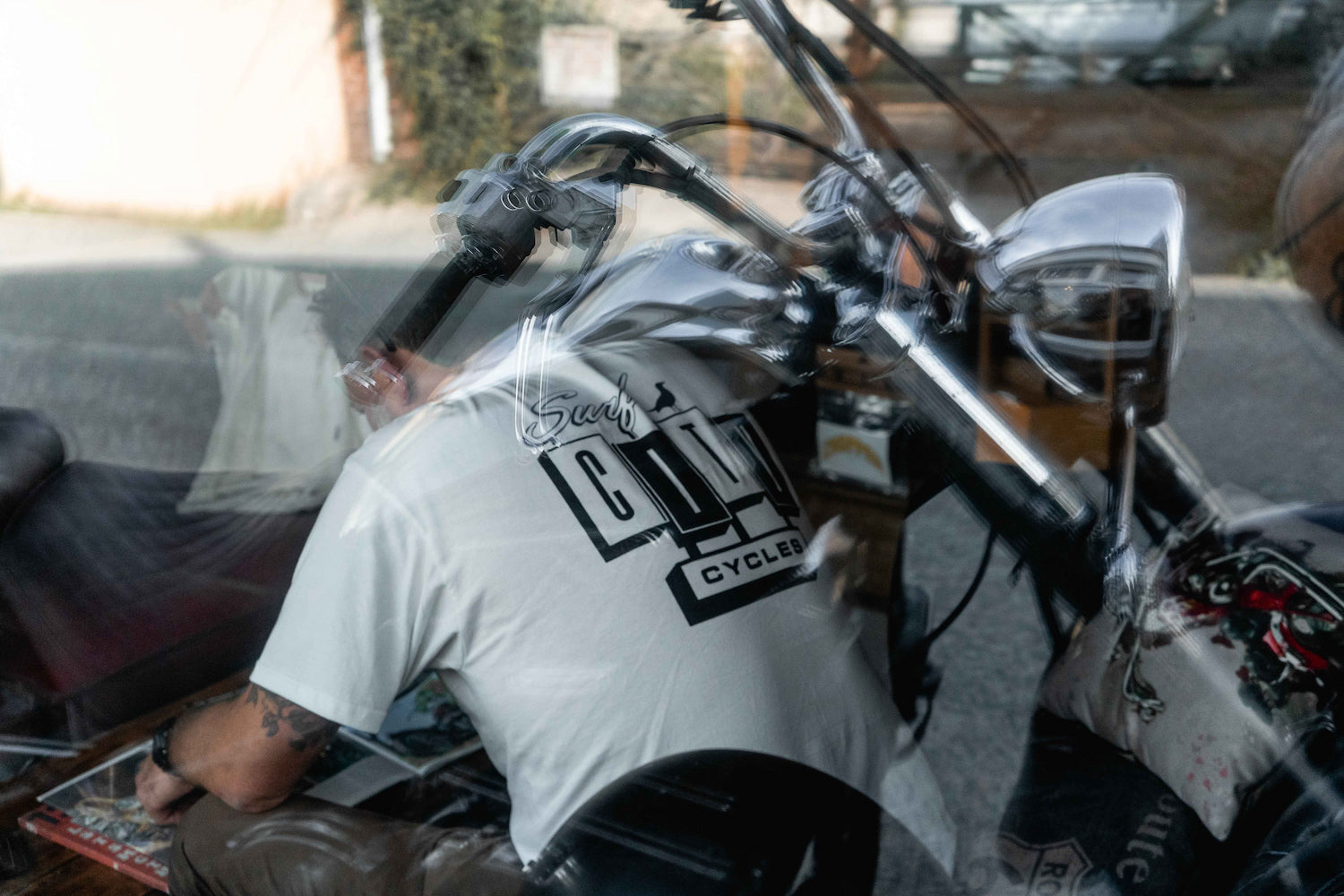 A double-exposure reflection of a man wearing a white OG Logo organic cotton tee looking over a motorcycle in the Surf City Cycles workshop.