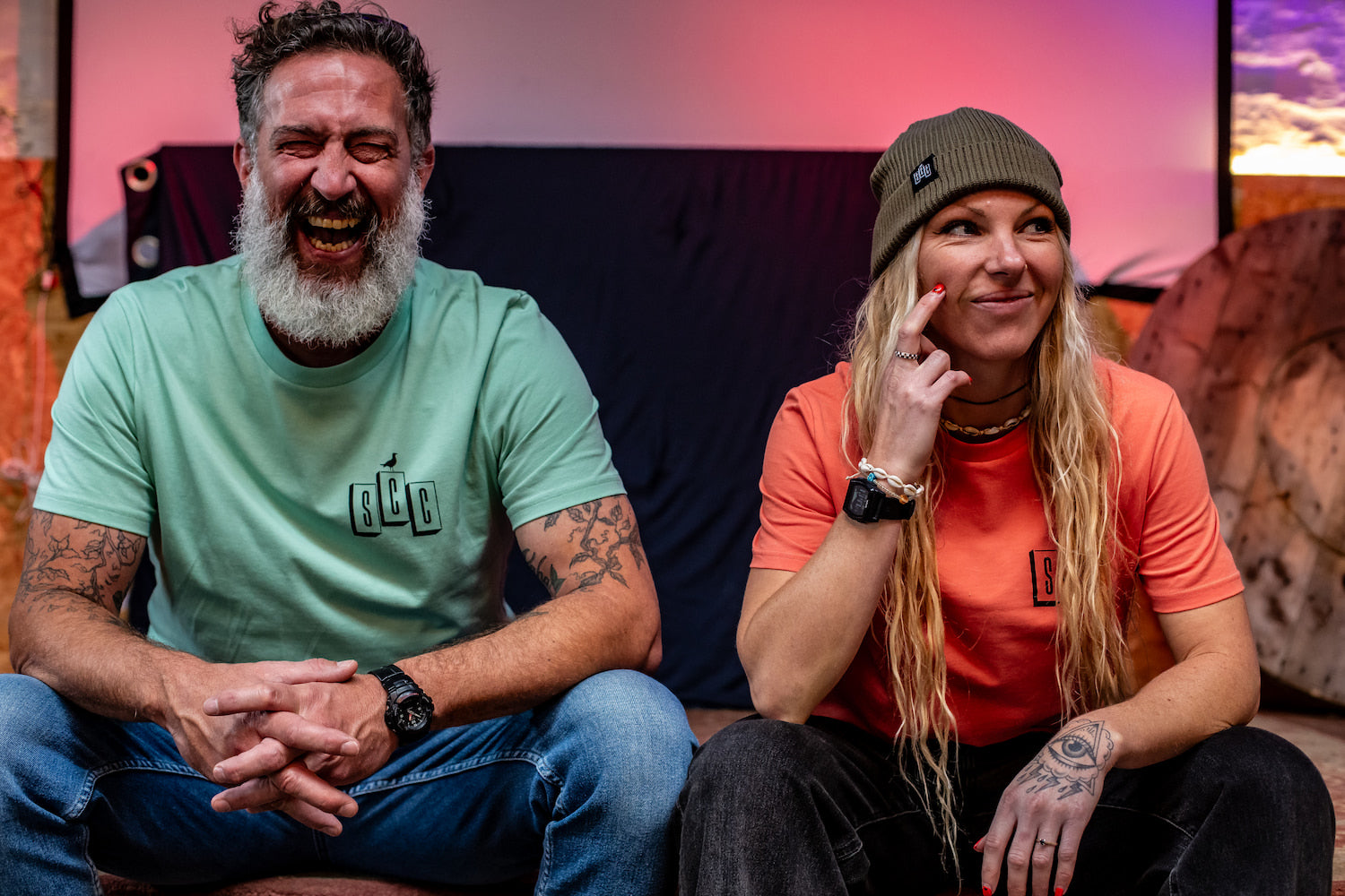 A man in a Seafoam OG Logo tee and a woman in a Fiesta coral tee and Olive ribbed beanie laughing together, showcasing the Surf City Cycles lifestyle.