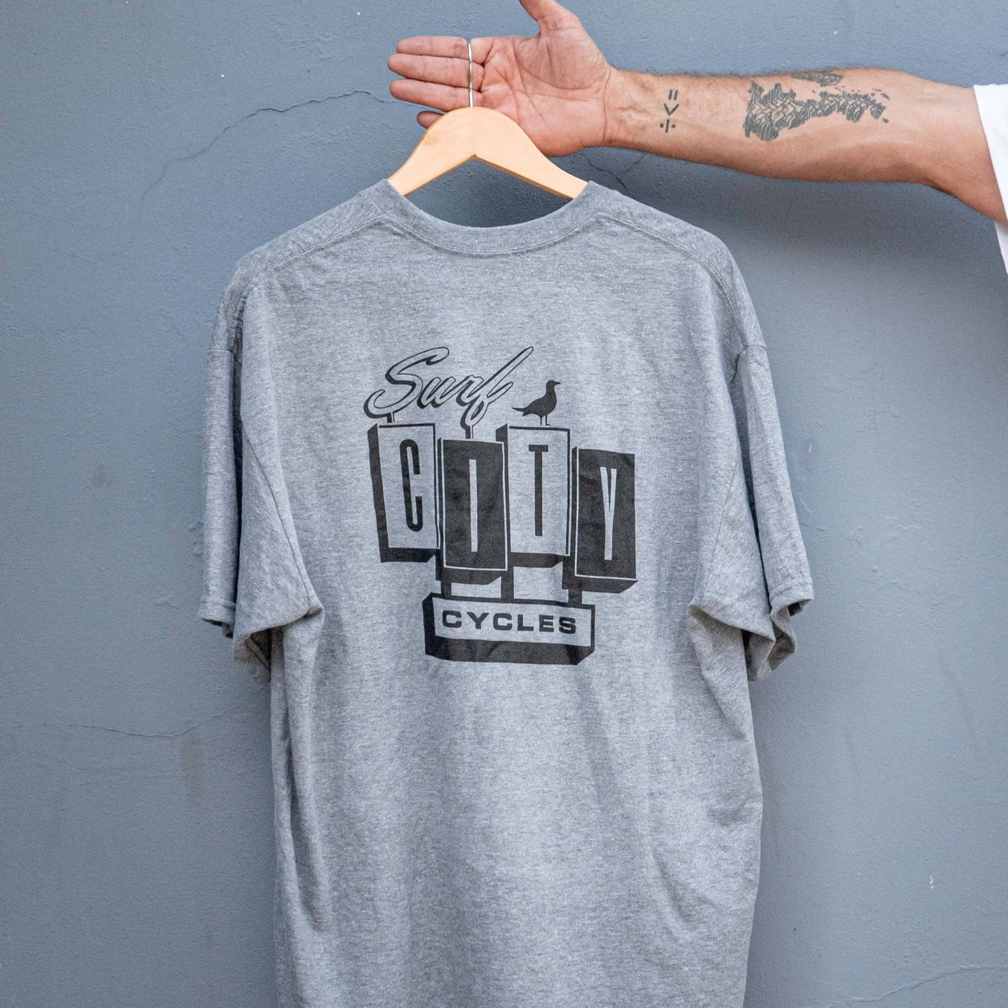 Rear view of the heather grey signature logo t-shirt on a hanger, featuring the large Surf City Cycles back print.