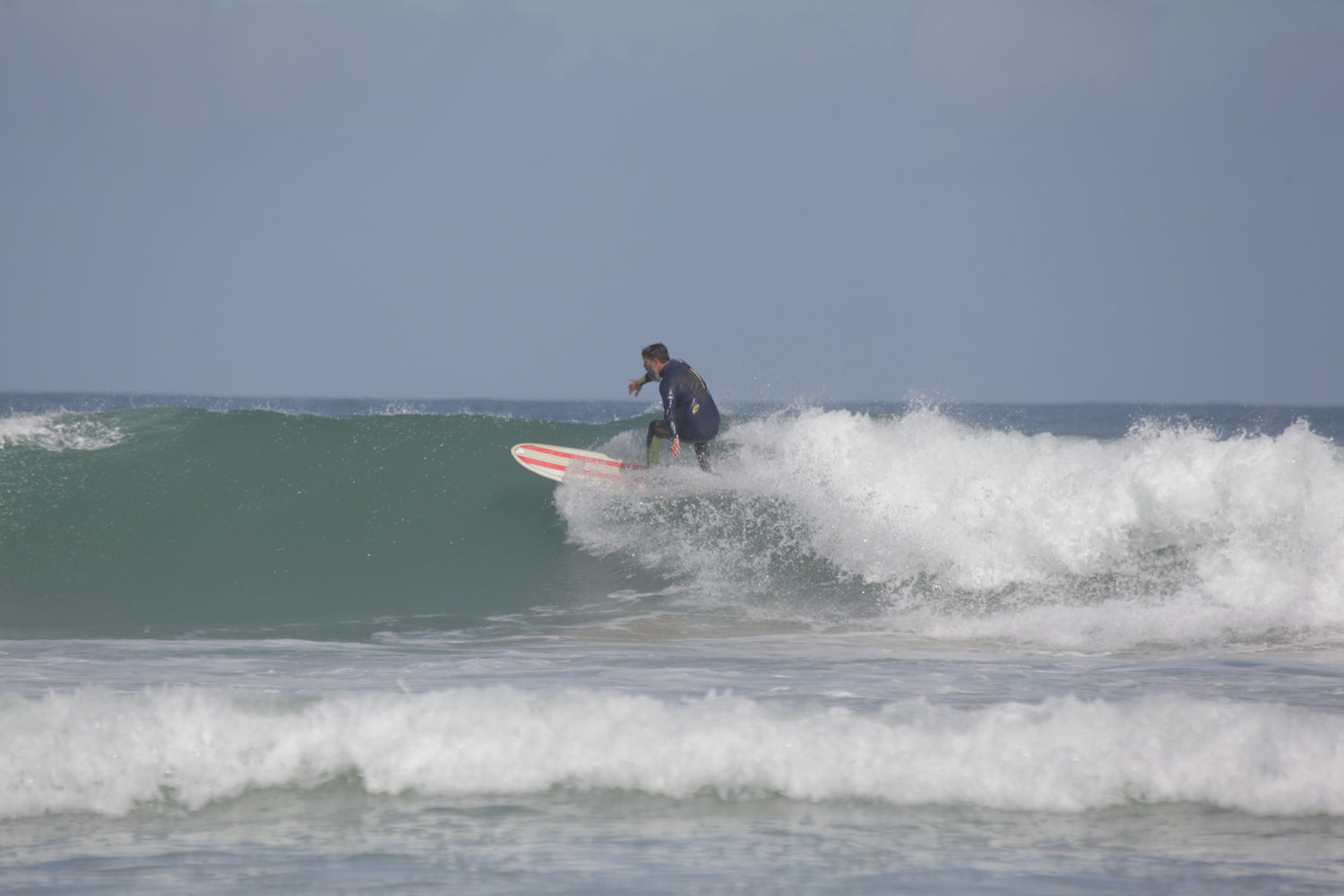 A man surfing a wave on a red and white striped surfboard in the clear coastal waters of Newquay