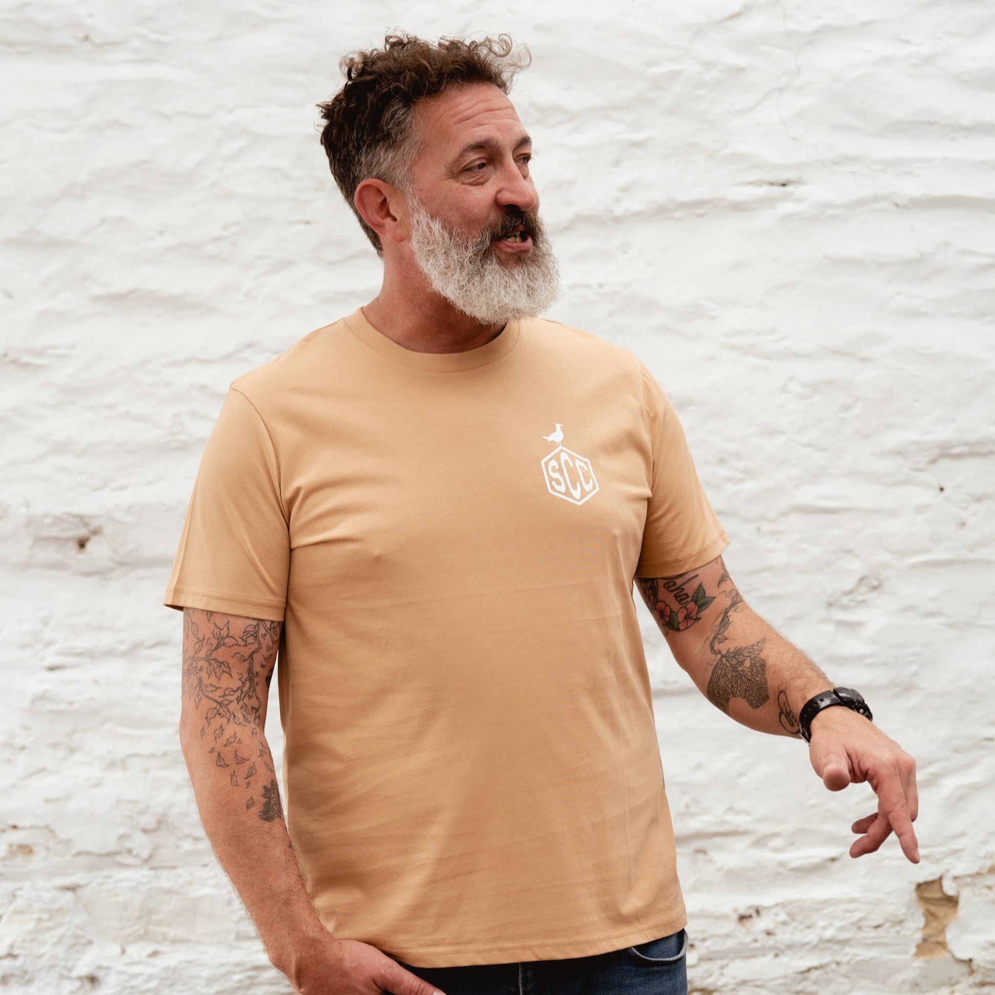 Side view of a man in a black SCC tee leaning against a workshop wall, capturing a rugged moto-lifestyle vibe.