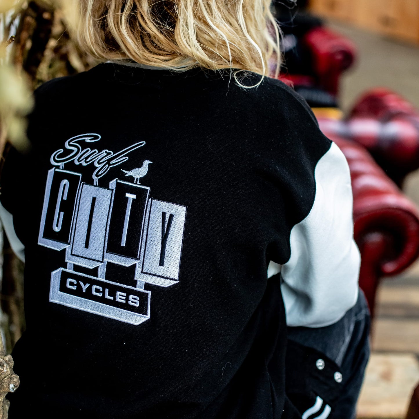 Rear view of the varsity jacket's back branding, showing the fit on a model with long blonde hair.
