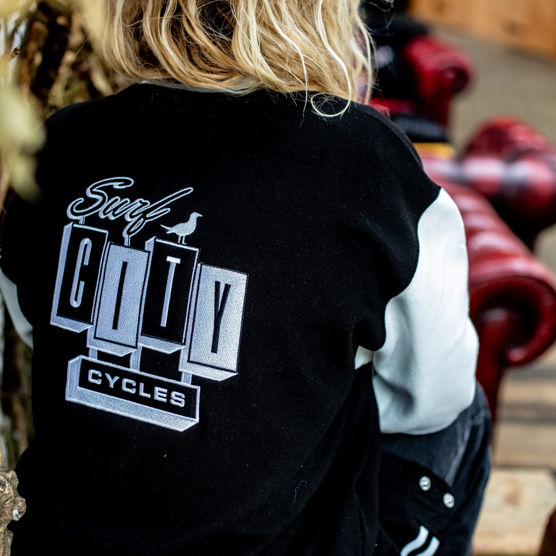 Rear view of the varsity jacket's back branding, showing the fit on a model with long blonde hair.