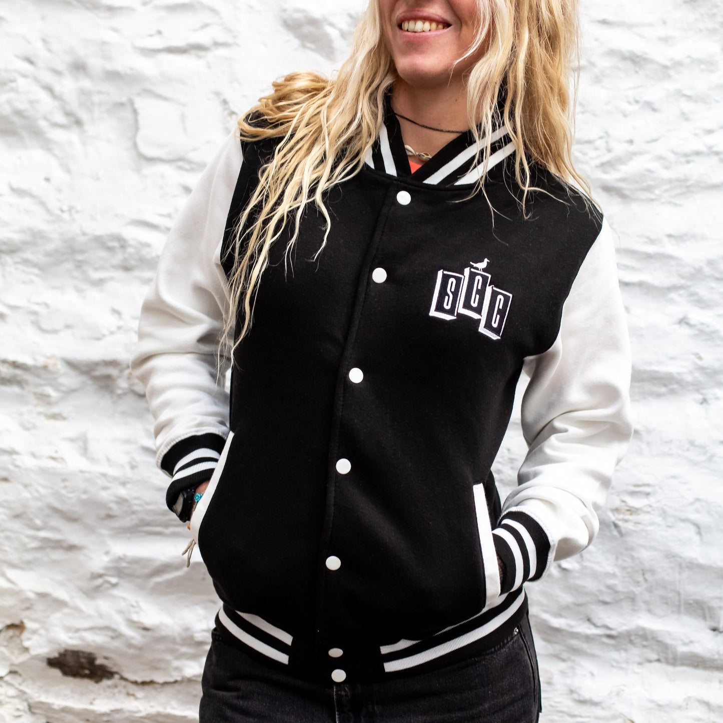 Female model smiling in the black and white varsity letterman jacket against a white brick background.