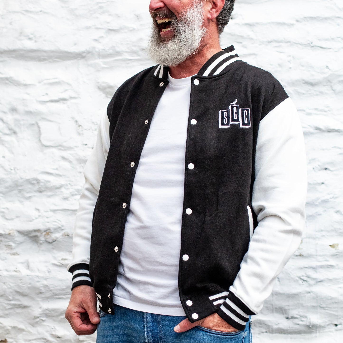 Bearded man laughing in a black and white varsity jacket with SCC embroidery over a white stone wall.