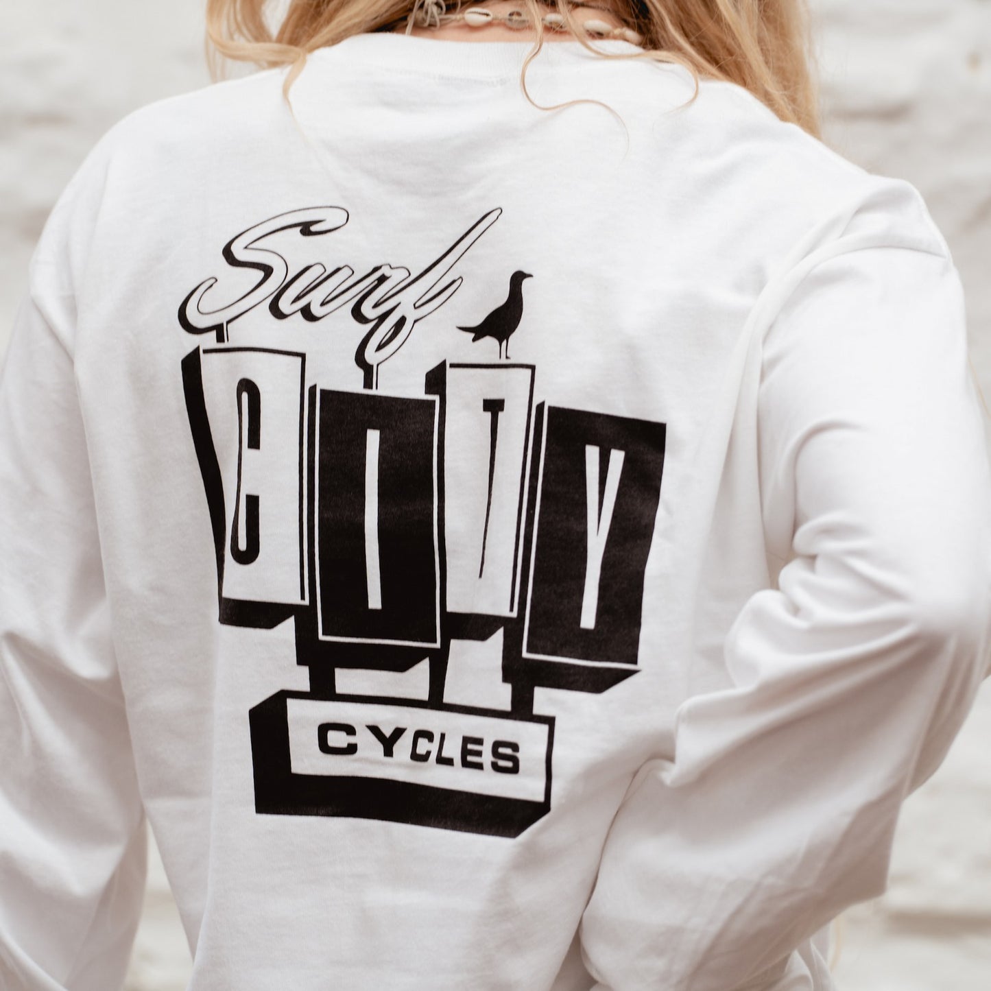 Lifestyle shot of a blonde surf girl in the white Surf City Cycles long sleeve tee leaning against a white stone wall in Project 83 Newquay