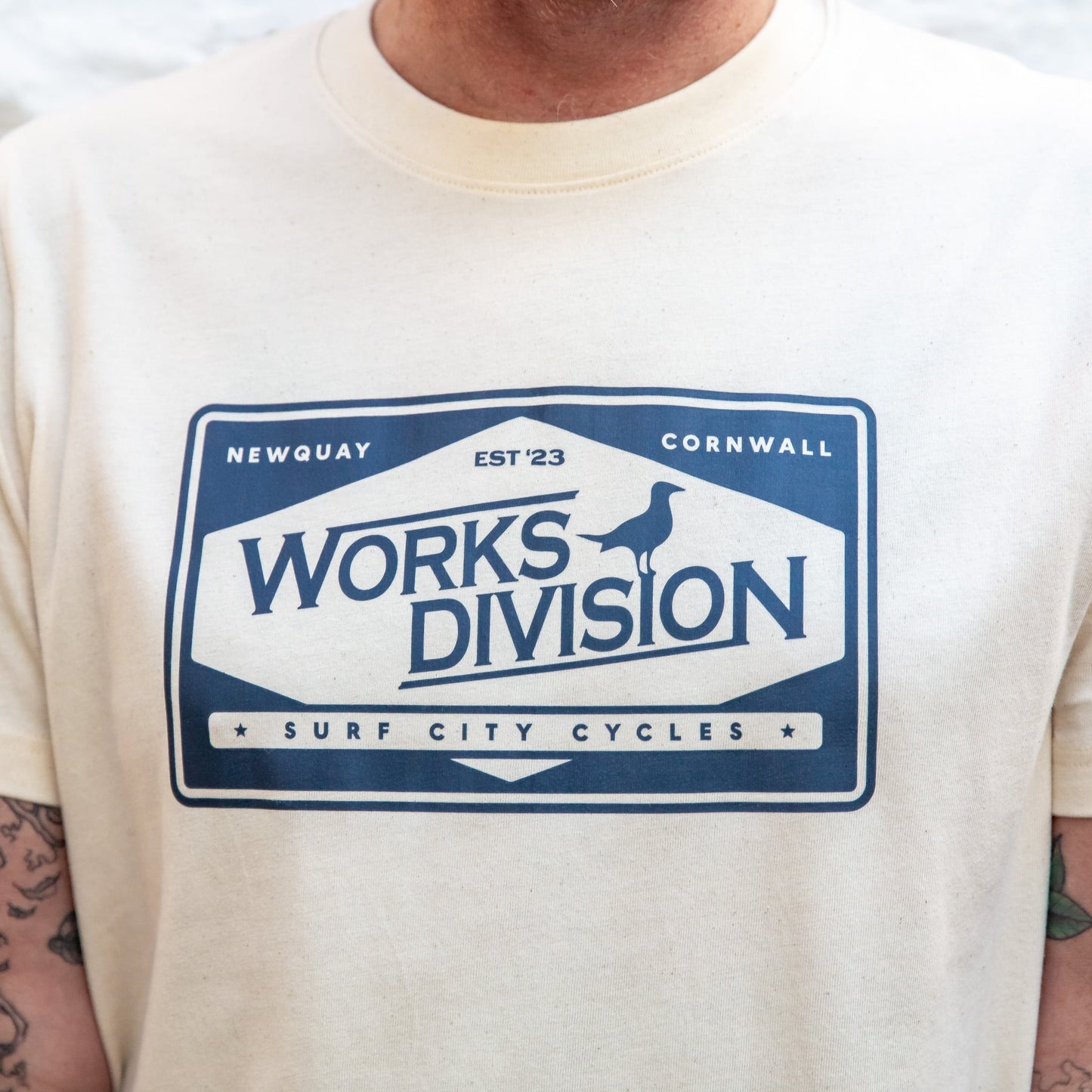 he Works Division Heavy Utility Tee in Natural, showing the relaxed unisex fit on a female model against a white stone wall.